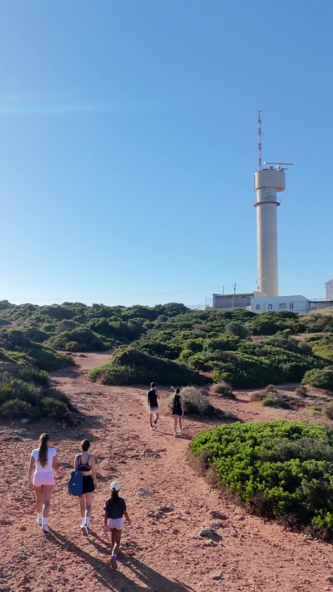 Caminhantes num trilho de terra vermelha rumo ao farol